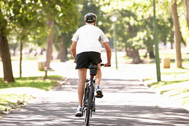 Rear View Of Mature Man Cycling Through Park Away From Camera During Camera